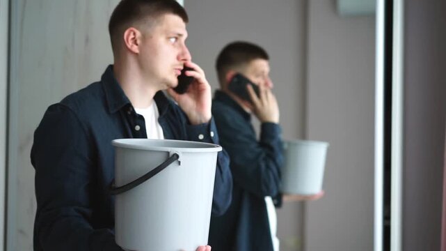 A man calls a roof repair service while collecting leaking water from the ceiling in a bucket.