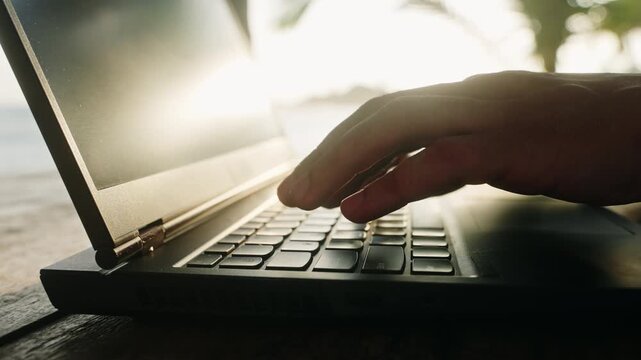 Man types on laptop keyboard at beach cafe, sunset backlight glows. Freelancer works remotely near sea, enjoys summer evening breeze, stays connected, meets deadline while travelling. Slow motion.