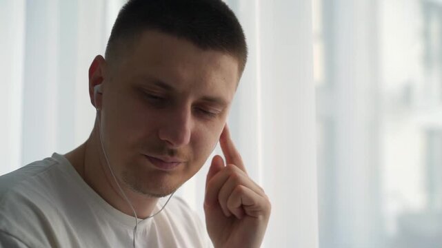 A young guy with a short haircut listens to music on headphones.
A man stands at a large window and listens to his favorite music.
Good mood guy