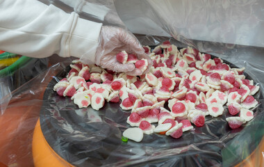 A gloved hand reaches into a pile of strawberry-shaped gummies on a factory floor.