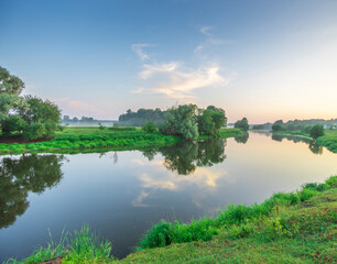 A picturesque river winds through a landscape of lush green fields and trees, reflecting the morning sky and clouds.