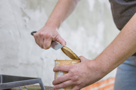 A man's hands use a utility knife to carefully pry open a can of paint, likely for a home renovation project.
