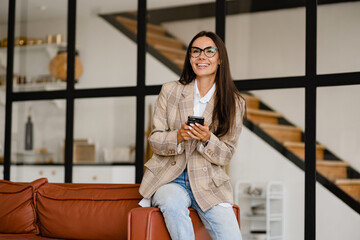 woman sitting on sofa in office relaxed using smartphone in beige jacket and jeans listening to earpods