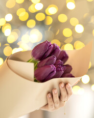 Bouquet of purple closed tulips in gift paper in hand on yellow bokeh effect background. Bouquet of flowers in festive romantic atmosphere. Floristics concept. Side view. High quality photo