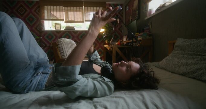 A woman uses a mobile phone lying on the bed. She is typing and taking selfies with a sad look