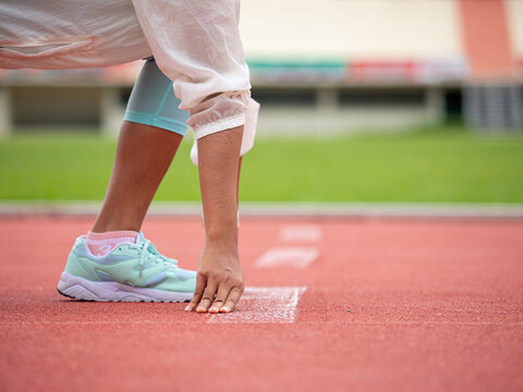Athlete in starting position on a track, wearing light blue running shoes, white socks, and white jacket, focused on hand placement before race