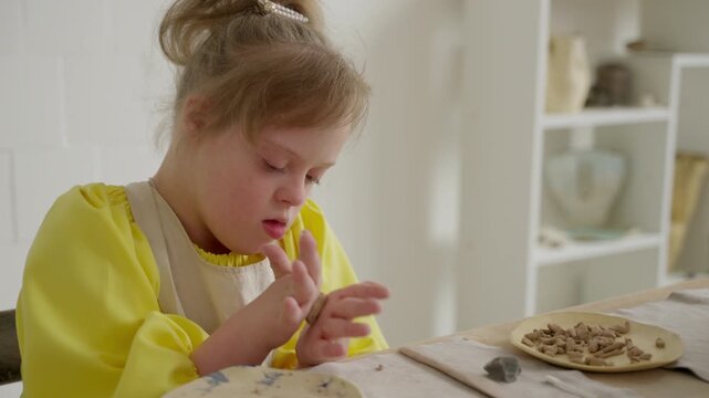Child with Down syndrome carefully works with clay at table in white-walled studio. Moment highlights importance of early art education in developing fine motor skills. Concept of creative learning
