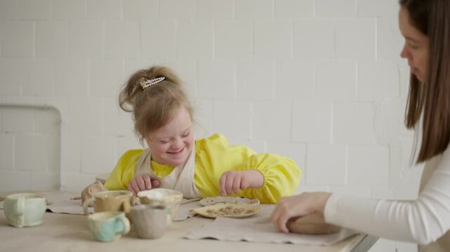 Laughing girl with Down syndrome and pottery teacher enjoy crafting in white-walled workshop. Scene showcases early educational support through guided creative activities. Concept of art education