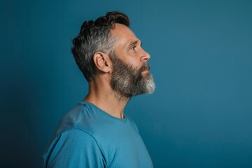 Fototapeta premium Profile portrait of middle-aged bearded man in blue t-shirt over blue studio background, copy space. Side view of handsome confident man posing on blue, standing straight and looking, Generative AI