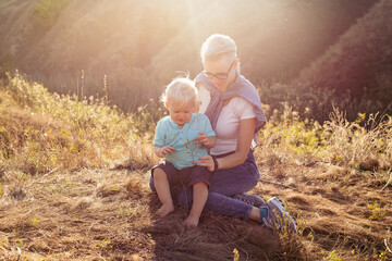 Baby boy and his mother in nature in summer sitting on grass with sun rays on sunset