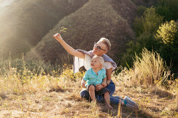 Mother and her son playing outdoors in nature in summer sitting on grass with sun rays on sunset