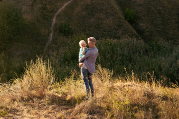 Mother holds her son in her arms and hugging in nature in summer with sun rays on sunset near green hills