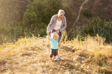 Little boy child takes his first steps and mother holds his hands in summer on grass with sun rays...