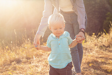 Little baby boy child takes his first steps and mother holds his hands in summer on grass with sun rays on sunset