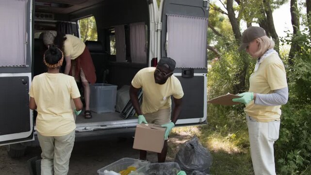 Full tilt-up shot of multiethnic team of cheerful young eco volunteers loading containers full of recycled rubbish into minivan trunk, event organizer checking report, while wrapping up forest cleanup