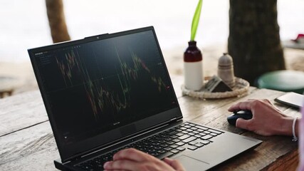 Crypto trader analyzes market graphs on laptop at beachside cafe. Sunset, mouse clicking, analyzing patterns, freelance finance work. Relaxed setting merges trading with leisure time at ocean shore. - Powered by Adobe