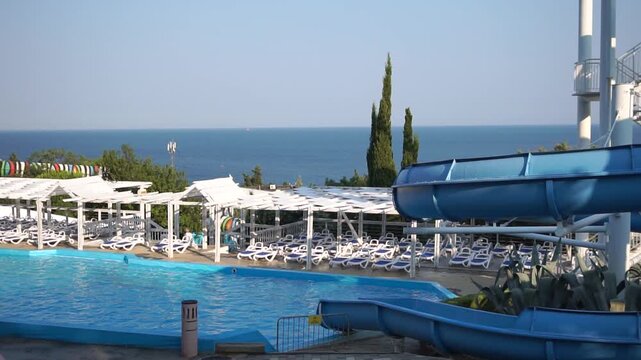 A blue waterslide curves down towards a pool, with rows of white lounge chairs and a view of the sea beyond.