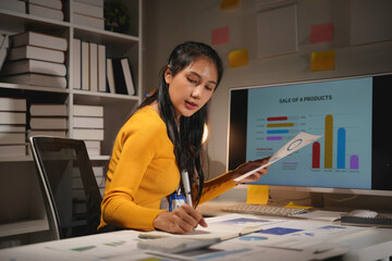 Young professional woman working overtime analyzing financial data and taking notes in her office at night