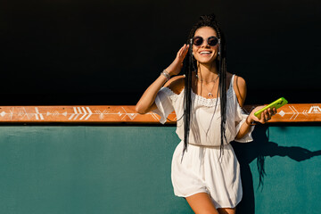 beautiful tanned smiling skinny woman posing on summer tropical vacation in white trendy dress using smartphone