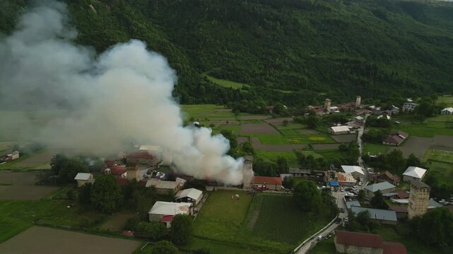 Aerial view of Svan tower in Mestia burning. Smoke rises in Svaneti mountains. Houses, green landscape visible. Emergency response, fire visible. Georgian scene with historical architecture. Drone