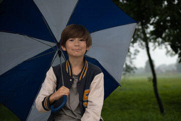 A young boy smiles brightly while holding a blue and white umbrella in a park. The background shows a misty, green landscape with trees and a hazy sky, indicating a rainy day. Copy space is available 