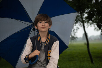 A young boy smiles brightly while holding a blue and white umbrella in a park. The background shows a misty, green landscape with trees and a hazy sky, indicating a rainy day. Copy space is available 