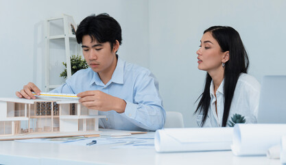 Fototapeta premium Professional male asian architect using ruler to measure house model length while young beautiful caucasian colleague using laptop to analyzed data on meeting table with house model. Immaculate.