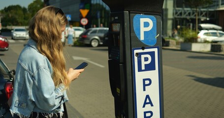Young beautiful woman in denim jacket stand at parking meter with smartphone. Female student with...