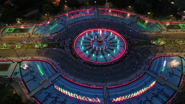 Aerial footage of the light show of the Bucharest fountains on a summer evening. Drone shot of the magnificent lights illuminating the mesmerizing dancing water fountain. Tourist attraction in summer.