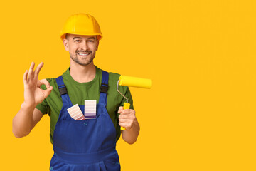 Portrait of male painter with color palettes and paint roller showing OK on yellow background