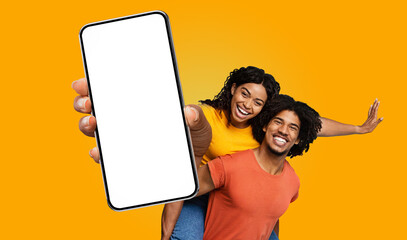 A young black woman in a yellow shirt is giving a piggyback ride to a man in a red shirt, both smiling broadly. The woman is pointing at the blank screen of a large smartphone