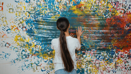Back view of young cute girl raise hand to paint the colorful stained wall. Attractive highschool student paint the wall with hand print while holding color palette. Creative activity. Edification.