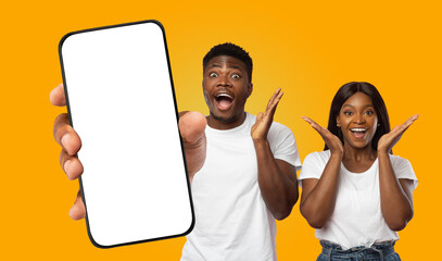 Black couple standing in front of a yellow background, looking at a blank smartphone screen being held out by a hand. Both have their hands raised in excitement with wide smiles on their faces.