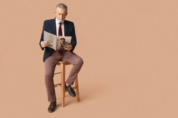 Fototapeta premium Handsome mature man in stylish formal suit with tie and magazine sitting on chair against beige background