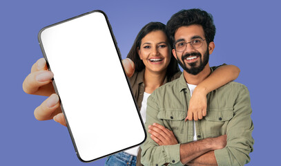 A young Indian couple is standing in front of a blue background, holding a smartphone with a blank...