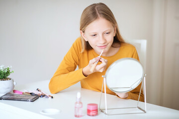 A teen girl with long blonde hair is sitting at a white table, applying lip gloss while looking in a mirror. She is wearing a yellow long-sleeved shirt.