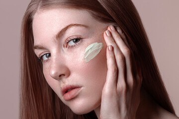 young beautiful natural no makeup red-haired woman applying green anti-redness cream on her face, skin care and sensitive skin treatment concept, beige background