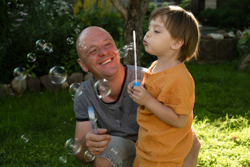 Fototapeta premium Dad and little son having fun on weekend blowing bubbles in backyard.