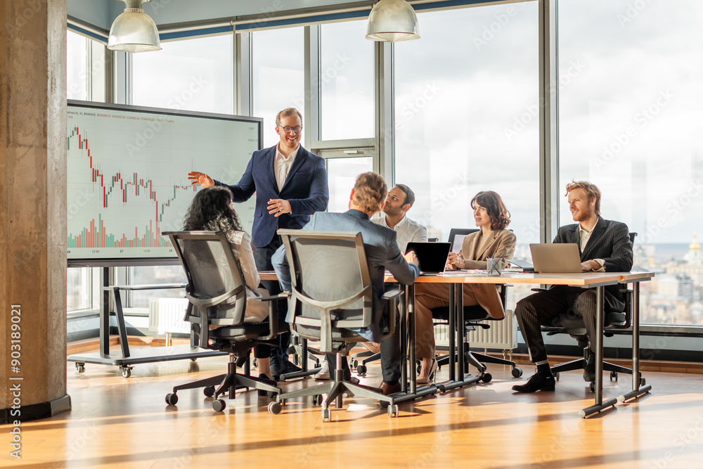 Poster A businessman stands in front of a large screen displaying financial data while presenting to a group of colleagues sitting around a table in a bright and modern office.