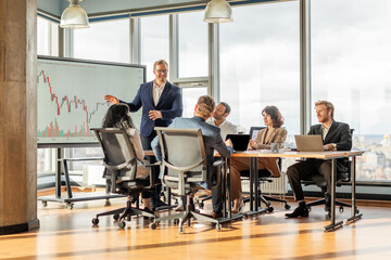 A businessman stands in front of a large screen displaying financial data while presenting to a...