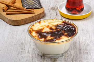 Traditional Turkish baked rice pudding called Fırında Sütlaç in glass bowl on white wooden board with Turkish tea and a cutting board with cinnamon sticks and a grater in the background