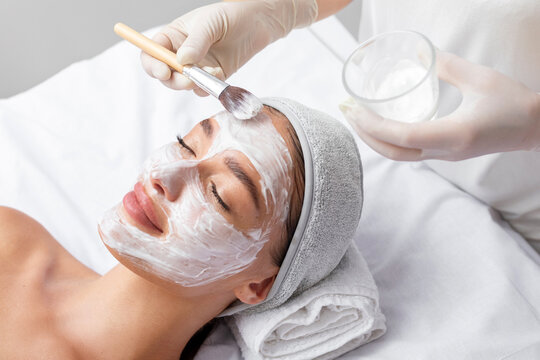 Beautician applying white clay mask to face of beautiful young woman in beauty salon. Spa treatments concept