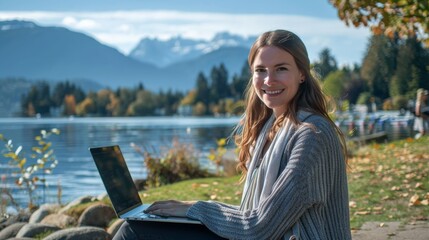 A young woman smiles as she sits on the grass beside a beautiful lake, working on her laptop, surrounded by fall foliage and mountains under a clear blue sky.