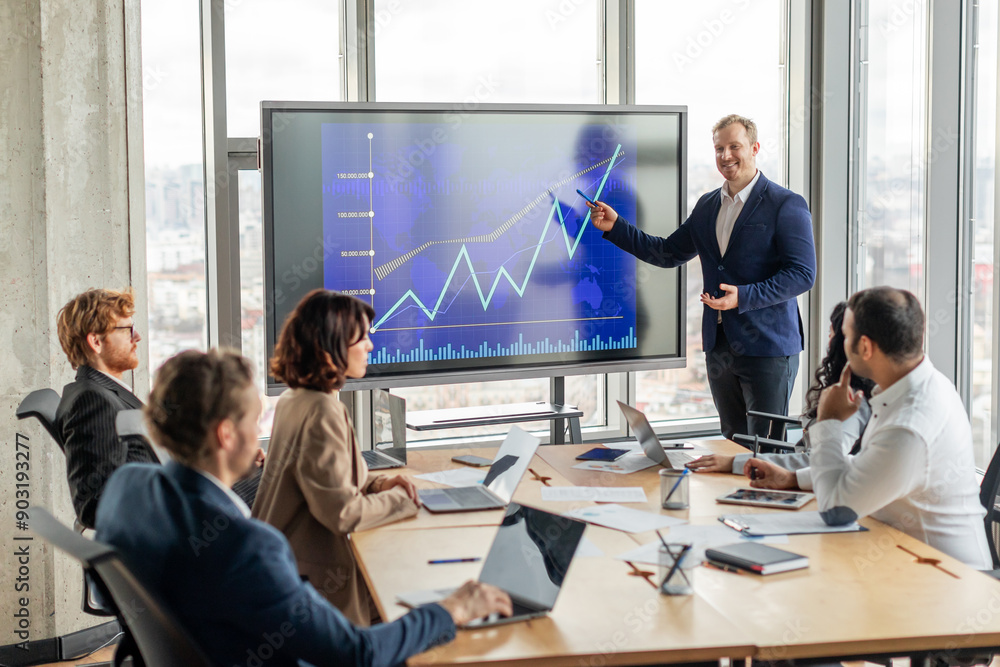 Fototapete A businessman stands in front of a large screen displaying a blue line graph that is trending upward, pointing to screen with a pen as he explains positive results to a group of attentive colleagues