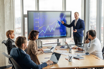 A businessman stands in front of a large screen displaying a blue line graph that is trending upward, pointing to screen with a pen as he explains positive results to a group of attentive colleagues