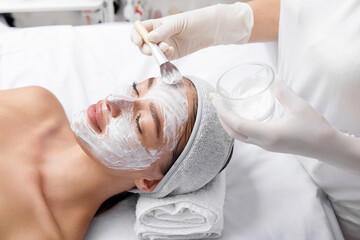 Professional cosmetologist applying nourishing facial mask on the patient's face. Moisturizing,...