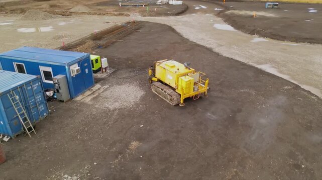 Aerial view of a compact tracked industrial machine operating beside a portable power unit and temporary site cabin, showing heavy-duty equipment used for construction, drilling or ground engineering 