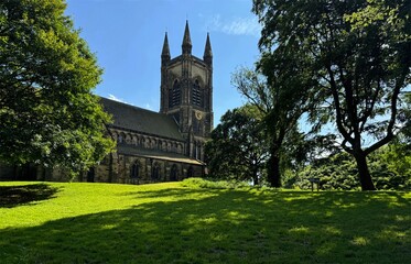 St Mary's grand Gothic-style church rises majestically among verdant trees, with sunlight filtering through to cast dappled shadows across the grassy lawn in Mirfield, Yorkshire, UK.