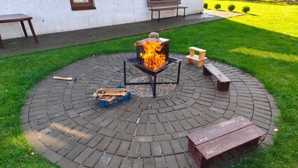 A metal fire pit is set up on a tiled area next to the lawn and benches. In it, a fire is burning...