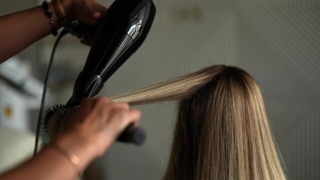 Close-up hands of unrecognizable female hairdresser drying and straightening long blonde client hair with comb and hairdryer in beauty salon. Concept of professional fashion hairstyling, slow motion.